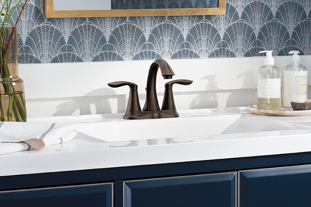 A dark bronze bathroom faucet installed on a white vanity countertop, with a white sink, blue cabinets, and patterned wallpaper in the background.