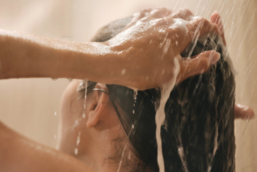 Close-up of a person washing dark hair under running water with soap lather, shown in a warm bathroom setting.