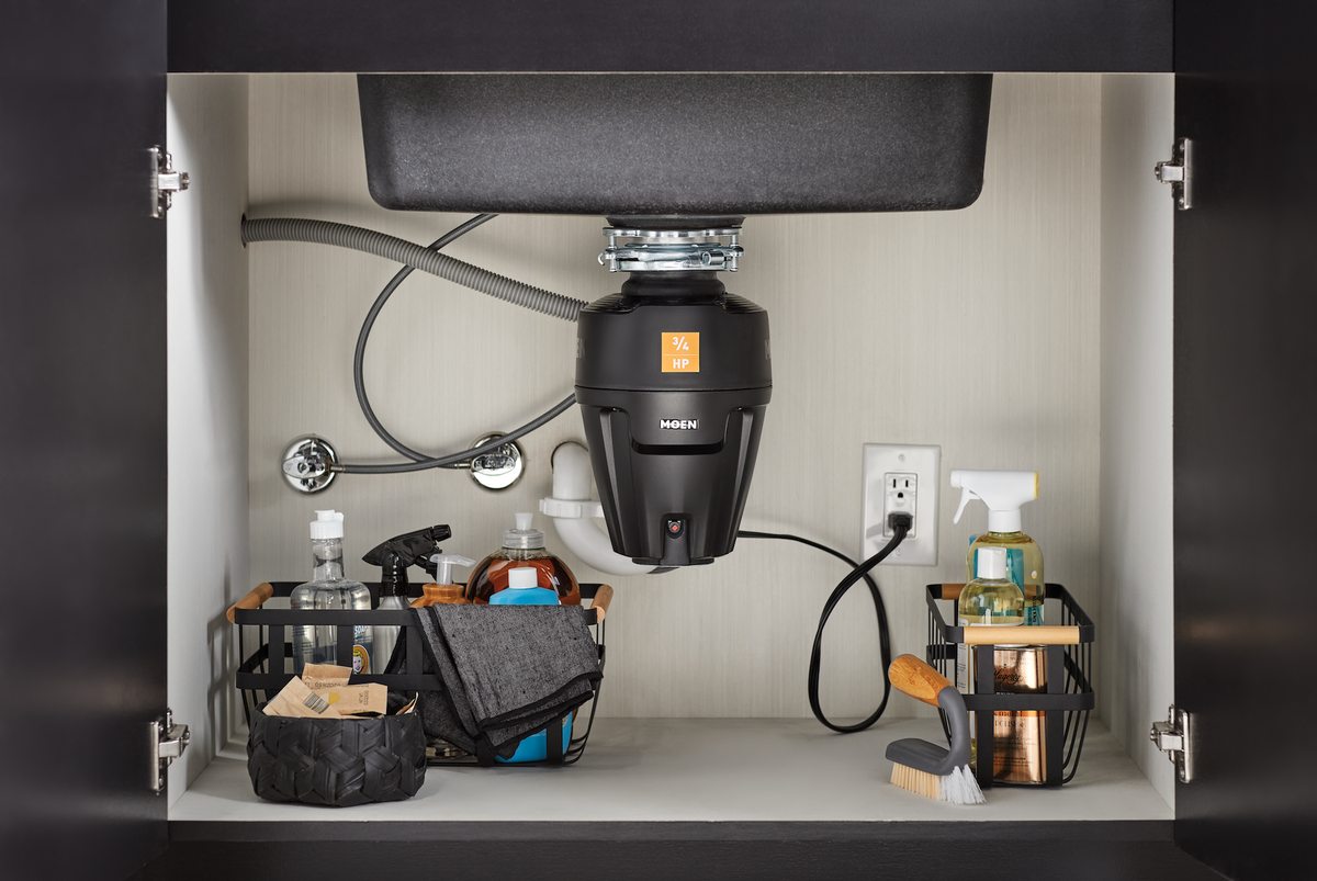 A black Moen undercounter garbage disposal installed in a white bathroom vanity cabinet, surrounded by kitchen sink accessories and cleaning supplies.