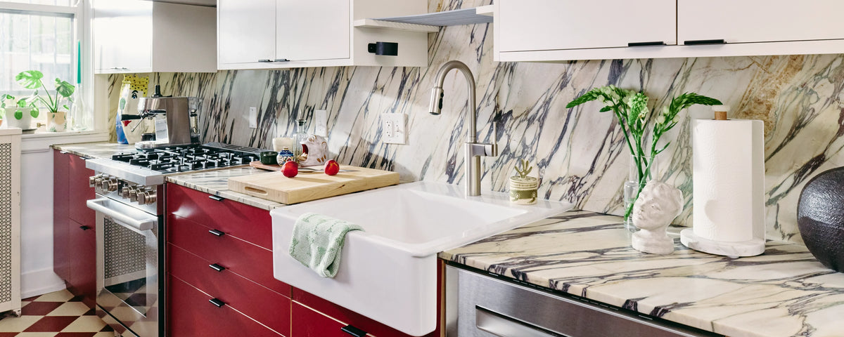 A modern kitchen interior featuring a white farmhouse sink, a high-arc gooseneck faucet, a white marble-look backsplash, and a white marble-look countertop with black veining. Red cabinetry, a gas stove, and kitchen accessories are visible.