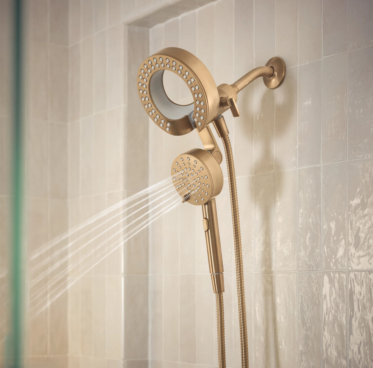 A wall-mounted shower system with a round overhead shower head, handheld shower head, and flexible hose, shown with water spraying from the handheld unit against a beige tiled wall.