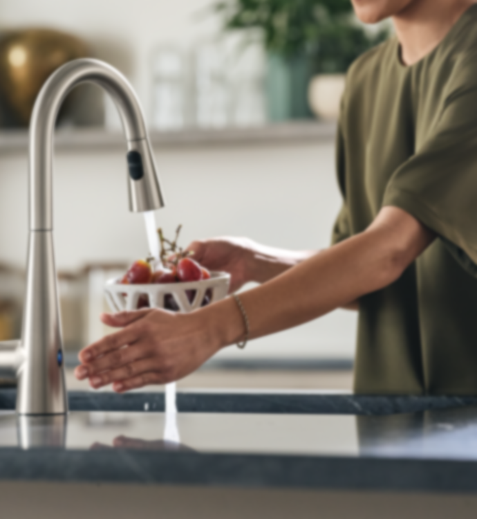 A modern stainless steel pull-down faucet with running water, shown in a kitchen setting with a person holding a white fruit basket under the stream.