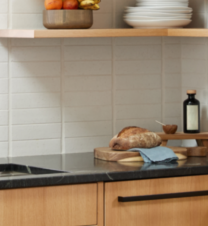 A close-up of a modern kitchen backsplash featuring light beige rectangular subway tiles, a dark stone countertop, light wood cabinetry, and open shelving with plates, fruit, and a bottle.