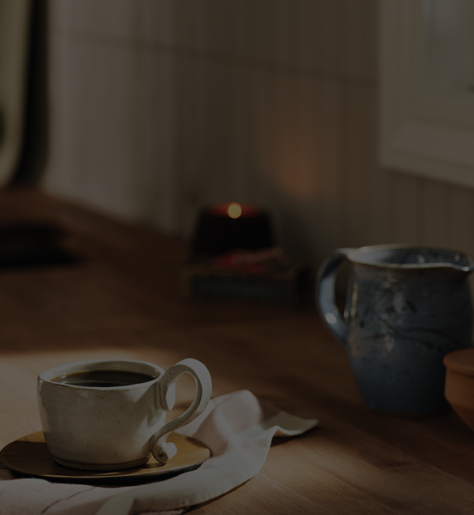 A rustic ceramic coffee cup filled with black coffee sits on a wooden table next to a blue stoneware pitcher, with a warm candle blurred in the background.