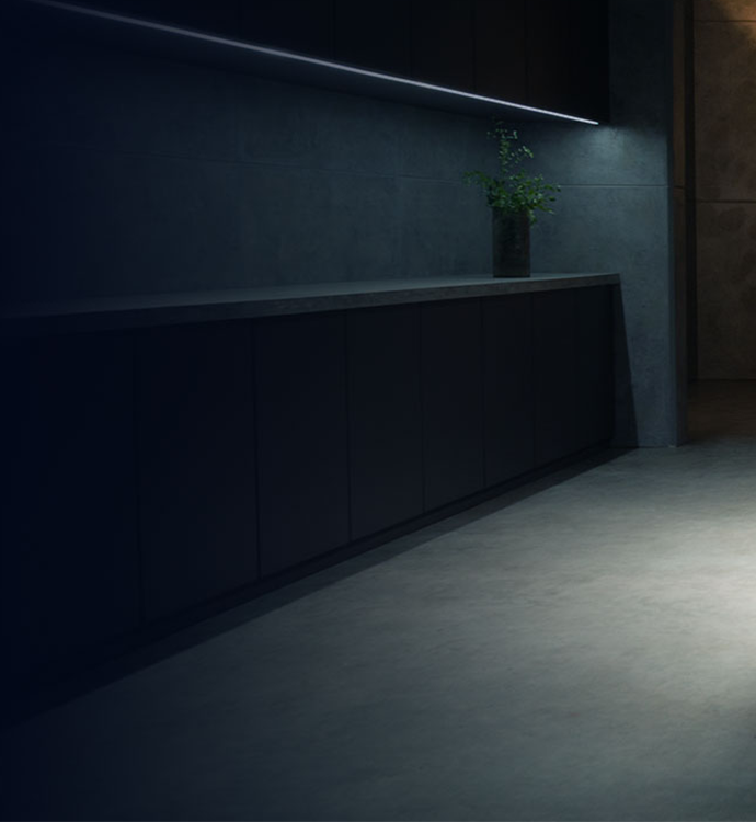 A modern dark wood kitchen cabinet with a matching countertop and integrated linear LED lighting, set against a dark gray tiled wall and concrete-look floor.