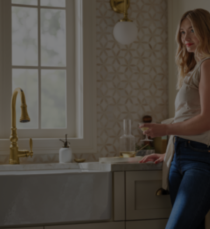 A woman in a beige sleeveless top and blue jeans stands in a kitchen next to a white farmhouse sink with a gold faucet, holding a glass in a softly lit interior.