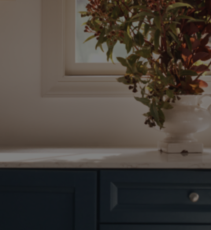 A close-up of a white countertop with a white pedestal vase holding leafy green and reddish-brown foliage, set against a warm beige wall and dark blue cabinetry below.