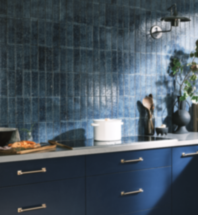 A modern kitchen scene with a blue tiled backsplash, matching blue lower cabinets, a light countertop, and small kitchen decor items including a white pot and wooden utensils.