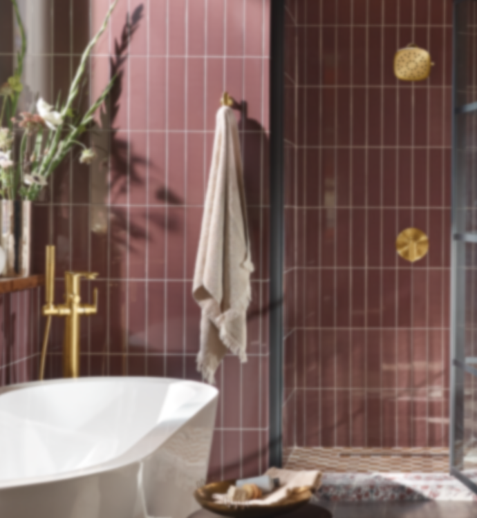 A modern bathroom scene with a freestanding white bathtub in the foreground, a beige towel hanging on a brass hook, and a walk-in shower with dark glass panels and gold-tone fixtures. The walls are covered in dark reddish-brown rectangular tiles.