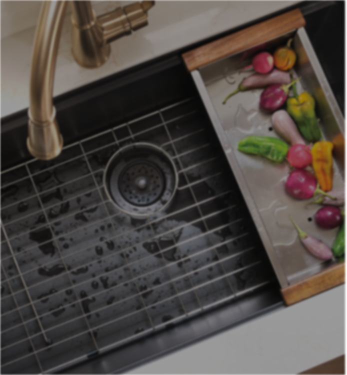 A stainless steel kitchen sink with a wire drain grid and a matching pull-out vegetable tray with wooden ends, shown in a real sink environment with a brass faucet and fresh vegetables inside the tray.