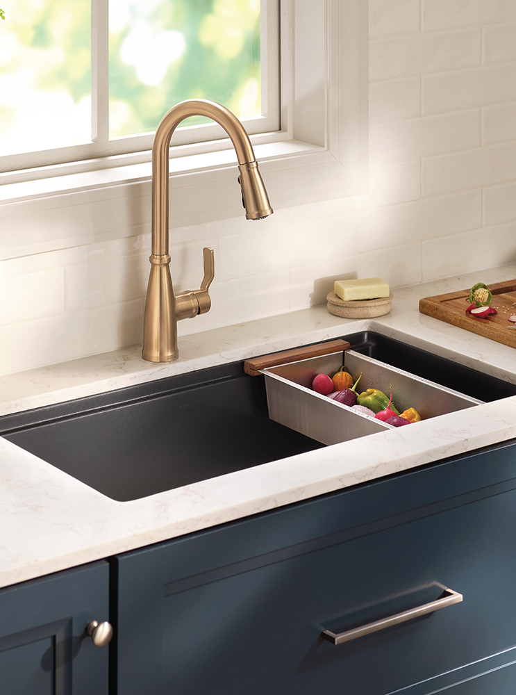A modern kitchen scene featuring a matte brass gooseneck faucet installed above a black undermount sink with a white marble countertop. A stainless steel divider tray holding colorful fruits sits in the sink, and a window with soft green outdoor light is visible in the background.