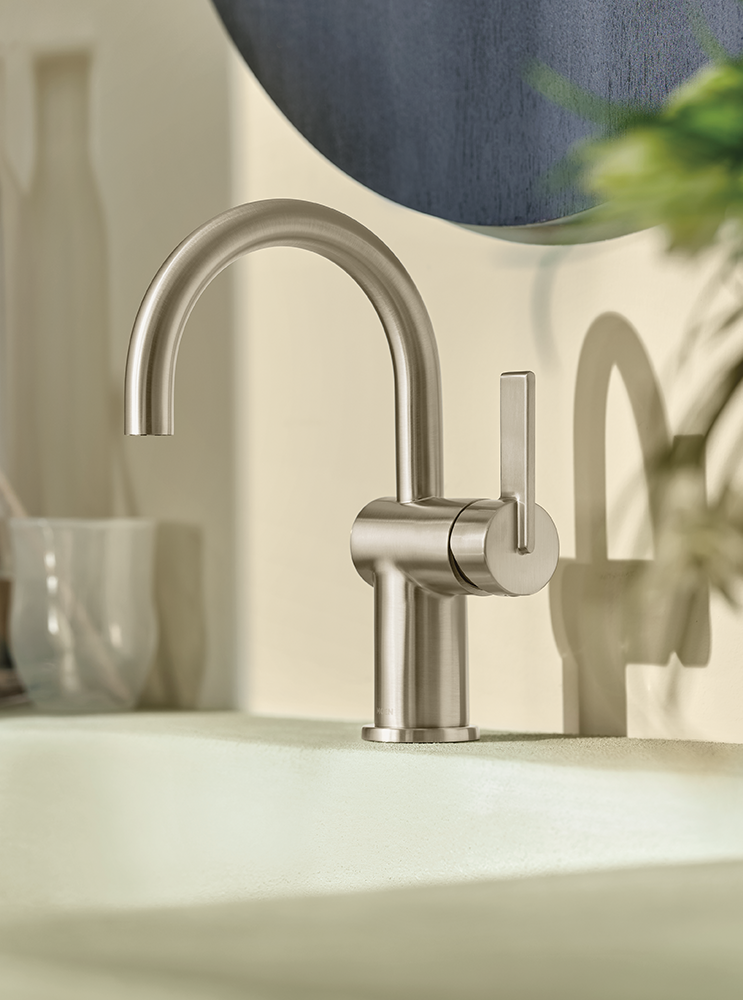 A modern single-handle bathroom faucet with a curved gooseneck spout and a vertical lever handle, shown installed on a sink in a softly lit bathroom setting.