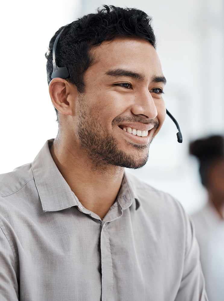 A smiling man wearing a light gray button-down shirt and a black headset, photographed in a bright office setting.