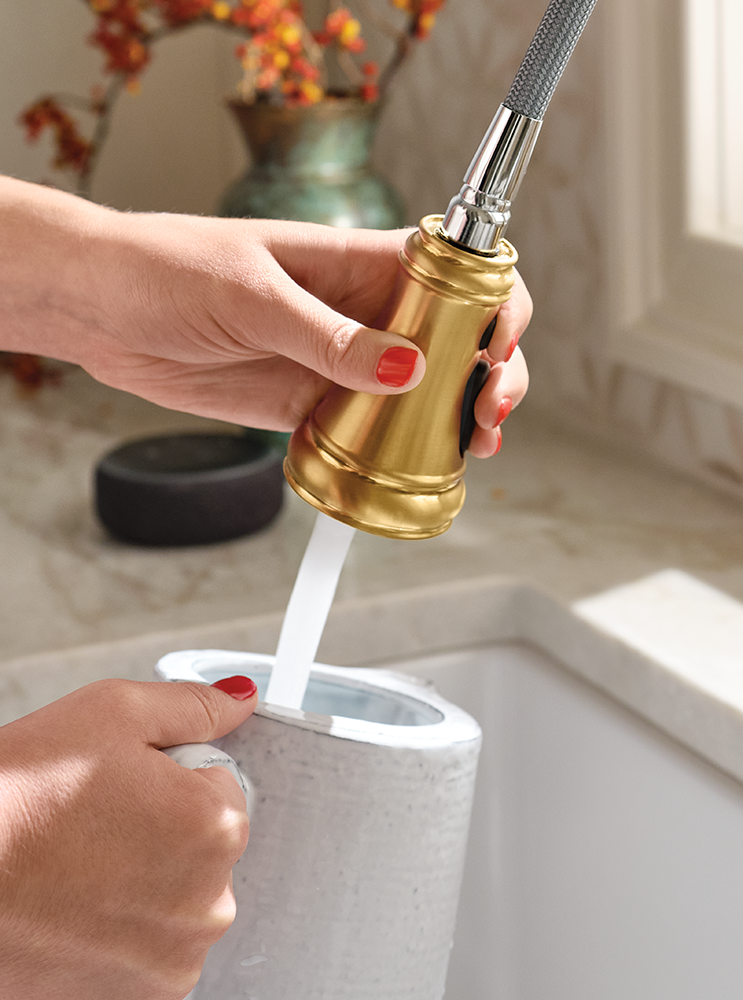 A close-up of a person's hands holding a gold and silver pull-out faucet sprayer over a sink, directing water into a white container.