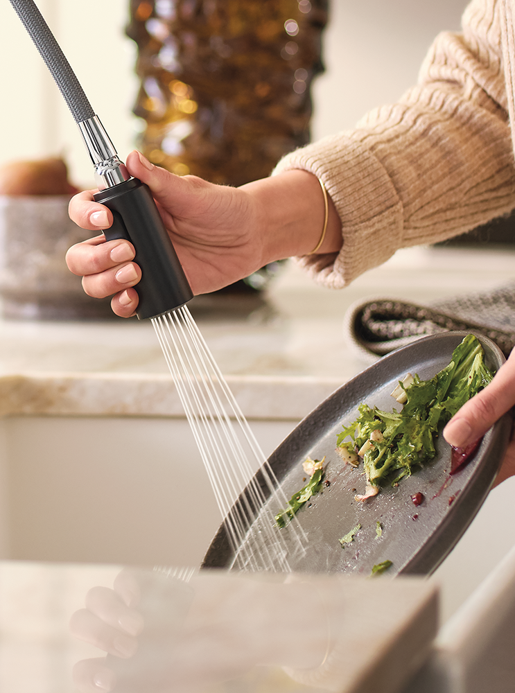 A close-up lifestyle shot of a person using a black pull-down kitchen faucet sprayer to rinse a dirty gray plate with running water in a kitchen sink.