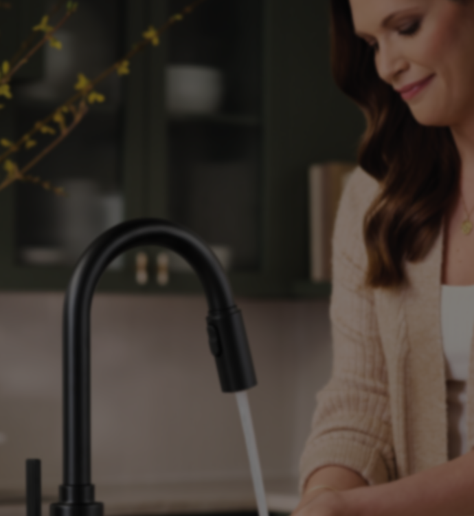 A woman in a beige cardigan stands at a kitchen sink using a matte black pull-down faucet with water running. The background shows dark green kitchen cabinets and a softly blurred interior.