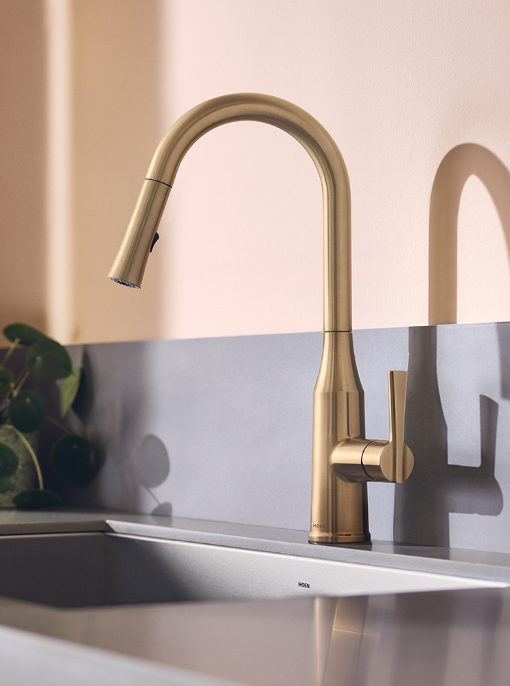 A modern single-handle kitchen faucet in brushed brass or gold finish, mounted on a stainless steel sink with a warm beige wall background.