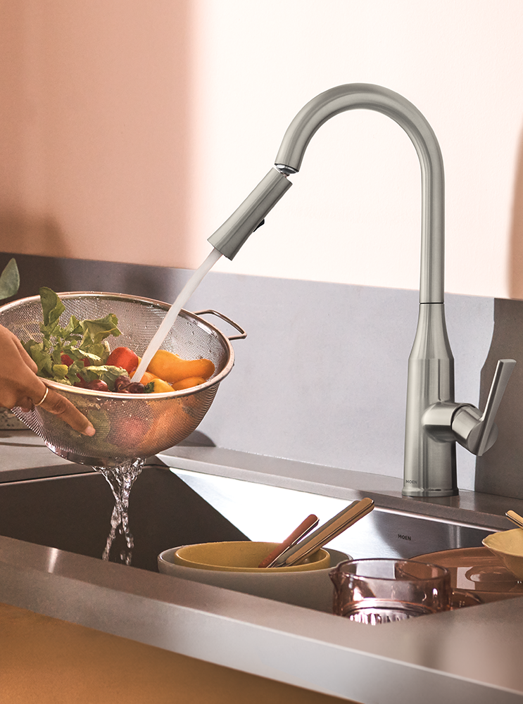 A modern pull-down kitchen faucet in a brushed silver finish, shown with water running from the sprayer into a stainless steel sink while a hand holds a metal colander of vegetables.