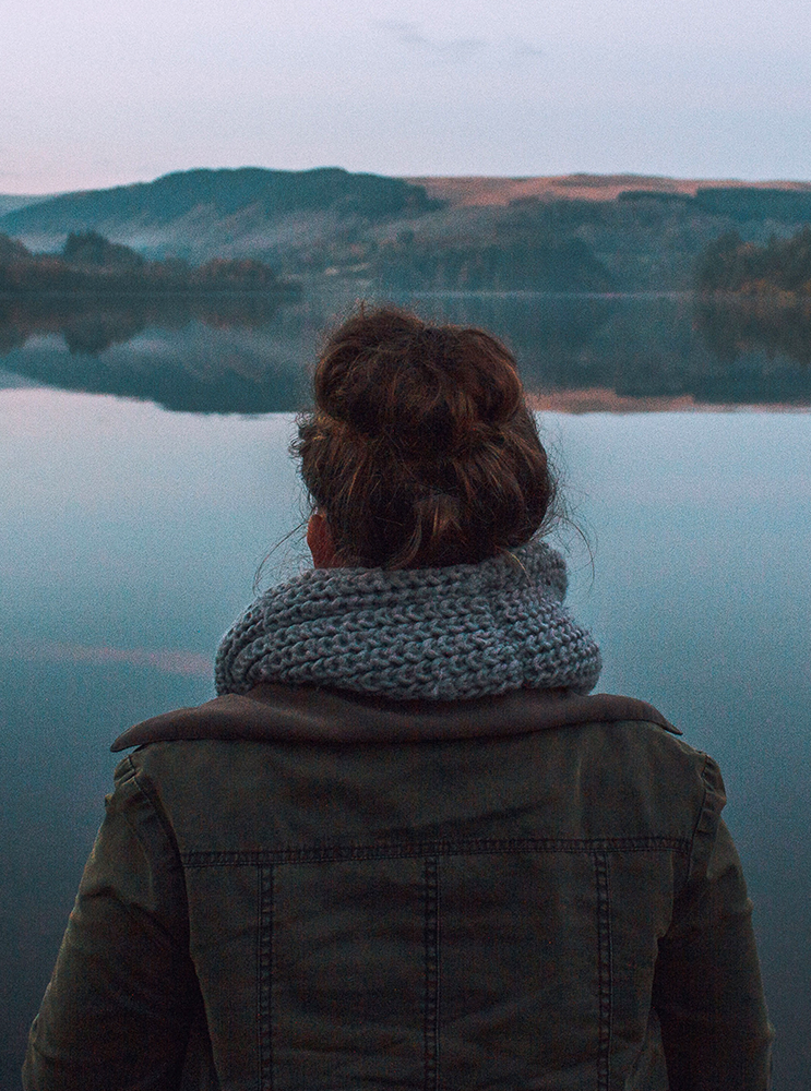 A person seen from behind standing by a calm lake with hills in the distance, wearing a dark jacket and a chunky knit gray scarf.