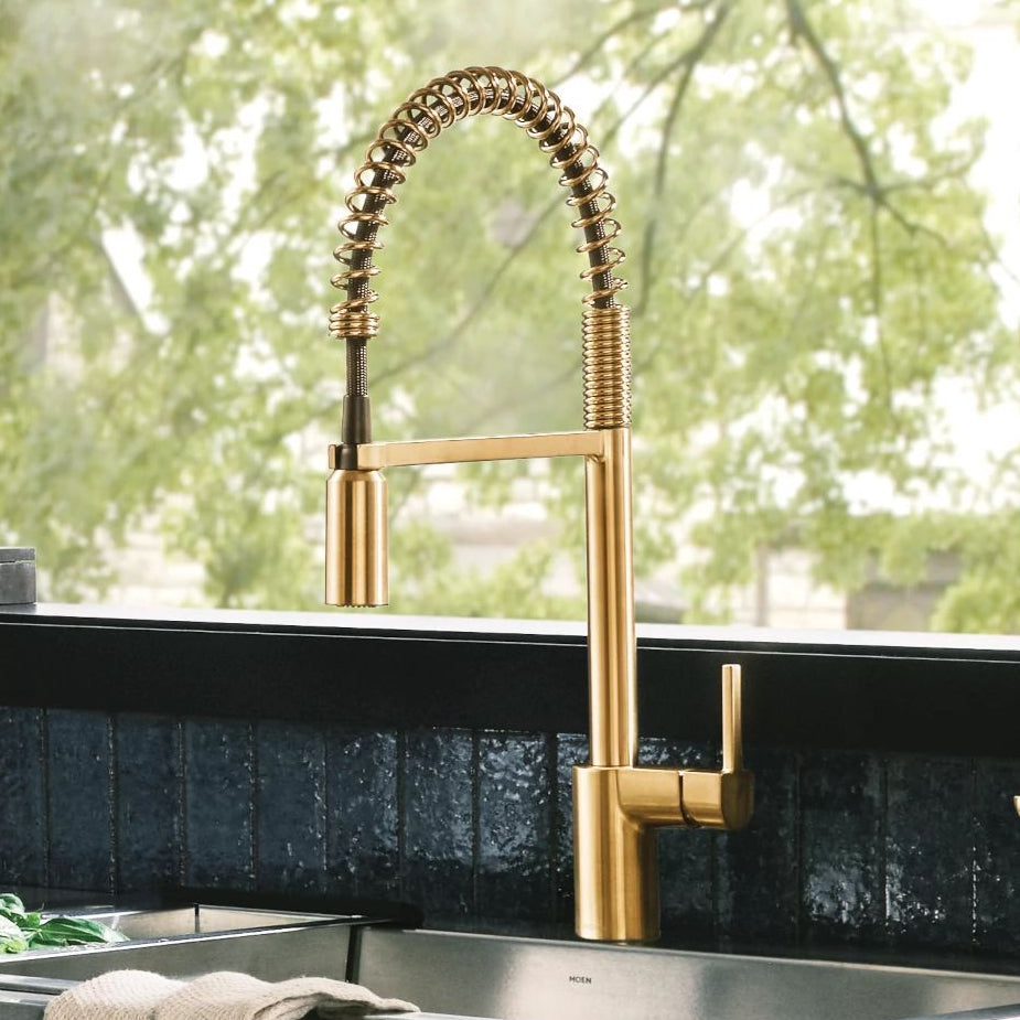 A modern kitchen sink setup featuring a tall, brushed gold pull-down faucet with a flexible spring-coiled neck, installed on a dark countertop with a matching secondary faucet partially visible on the right.
