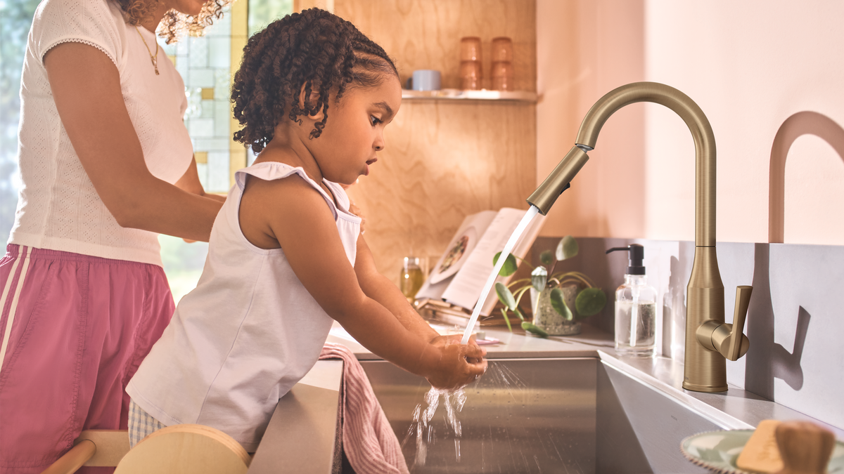 A child washes their hands under running water from a modern matte bronze faucet while an adult stands behind them in a bright kitchen setting.