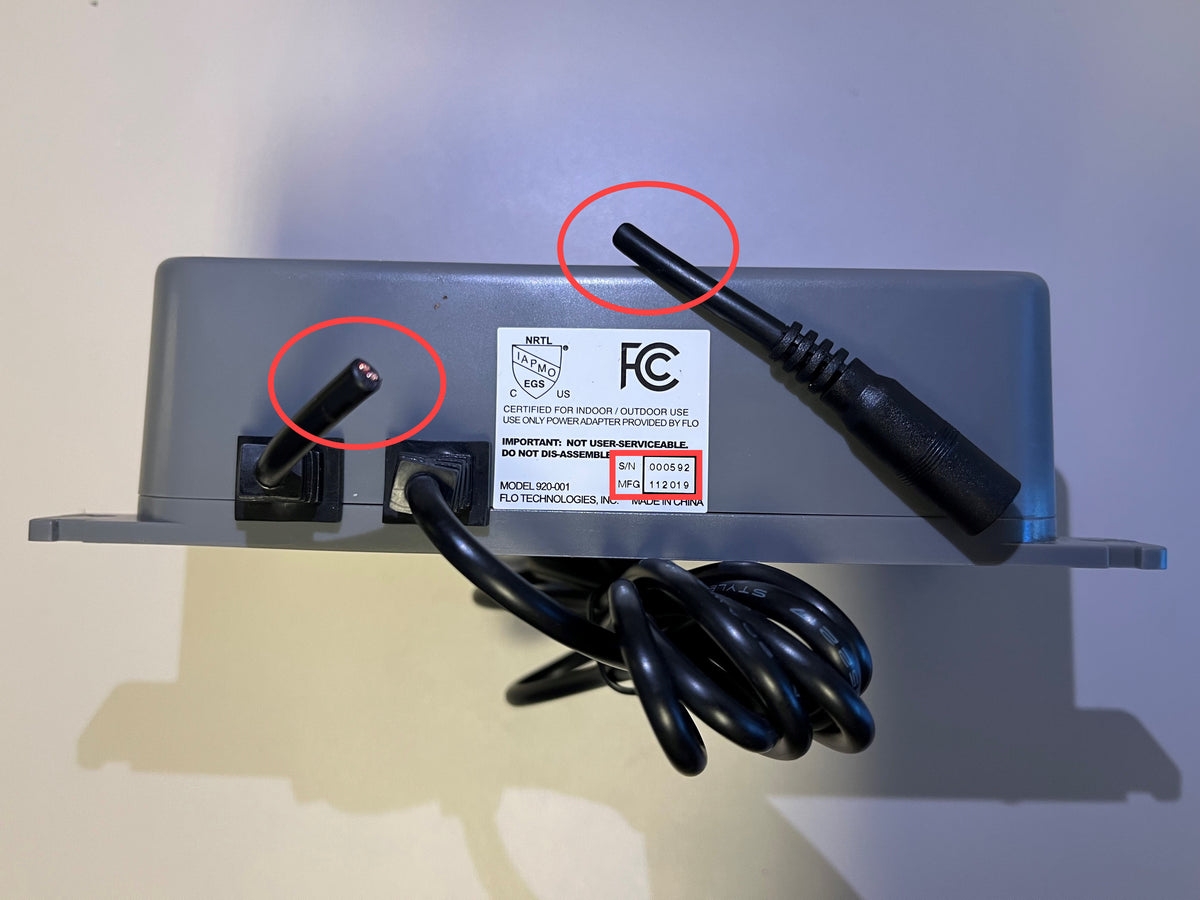 A close-up of the back of a gray electronic device showing its power adapter and cable, with two circular red marks highlighting the connector details.