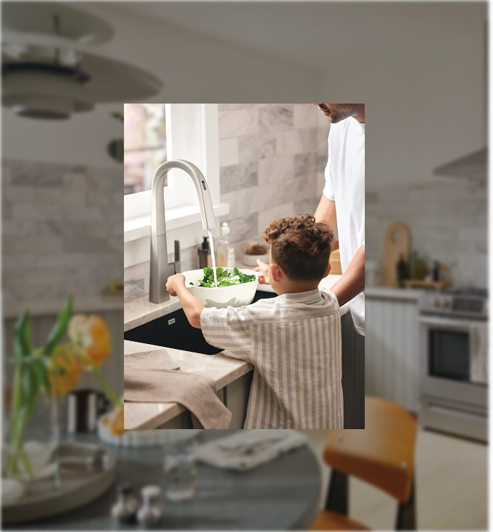 A lifestyle scene showing a young boy in a striped shirt washing vegetables at a kitchen sink while an adult man assists him. The image is presented as a centered square overlay on a blurred background of a modern kitchen.