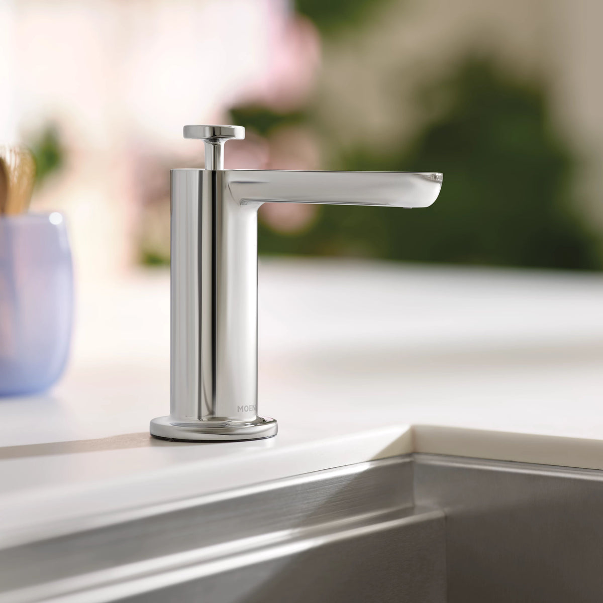 A modern chrome soap dispenser faucet with a sleek cylindrical body and angled spout, standing on a white countertop next to a stainless steel sink basin.