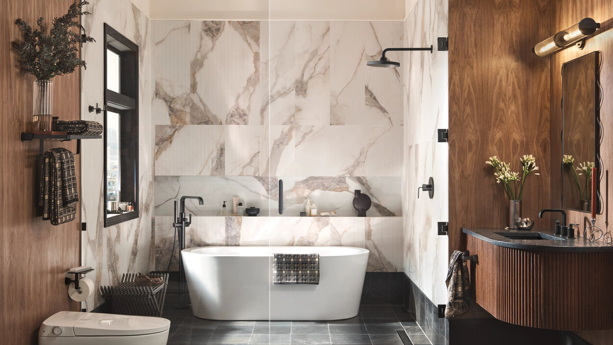 A luxury modern bathroom featuring a freestanding white bathtub set against a large marble wall, with dark wood paneling, a dark vanity, and a toilet paper holder on the left.
