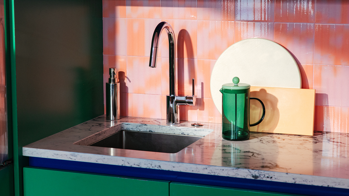 A modern kitchen sink setup with a tall chrome faucet, a marble-look countertop, a green French press, and a round white plate against a pink-orange tiled backsplash.