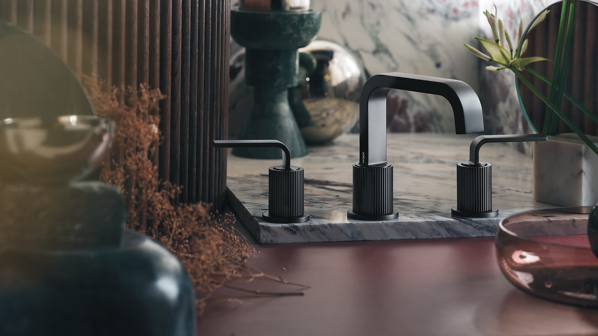 A modern three-hole bathroom faucet set in matte black, mounted on a gray marble sink deck with matching black cylindrical handles and spout, shown in a styled bathroom setting with warm lighting and decorative objects.
