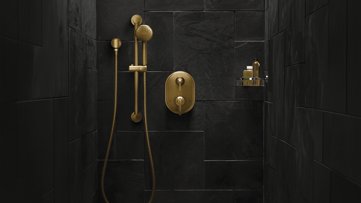 Gold shower fixture set installed on a black tiled wall, including a handheld showerhead with hose, round control plate, and small wall-mounted bottle holder.