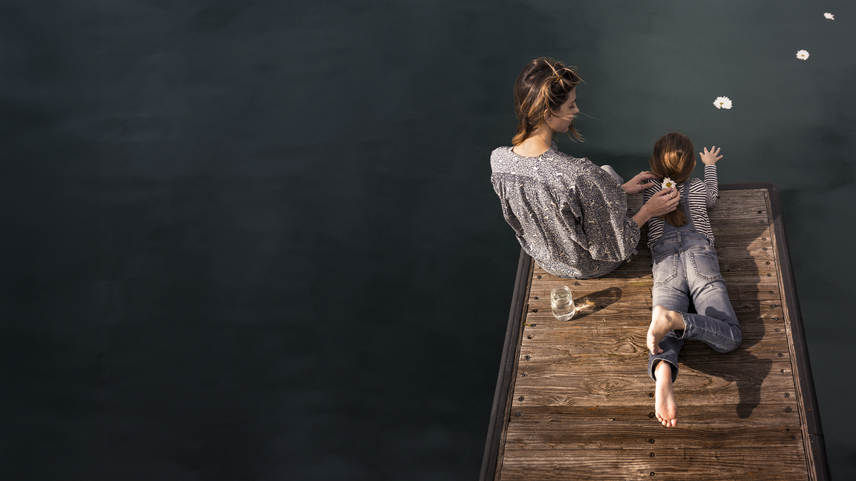 A woman and a young girl sit on a wooden dock over dark water, viewed from above. The woman wears a gray patterned top, and the girl wears a matching gray outfit.
