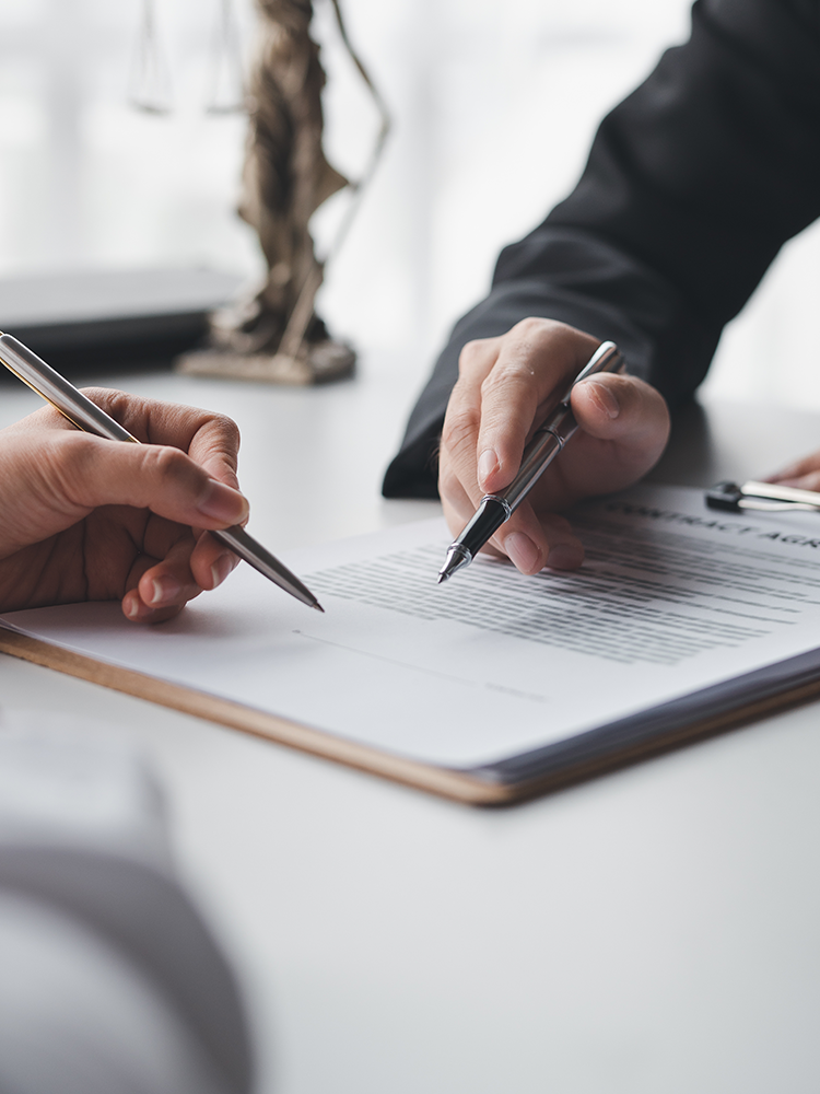 Two hands holding silver pens, pointing at a document on a clipboard over a white desk, with a blurred office background.