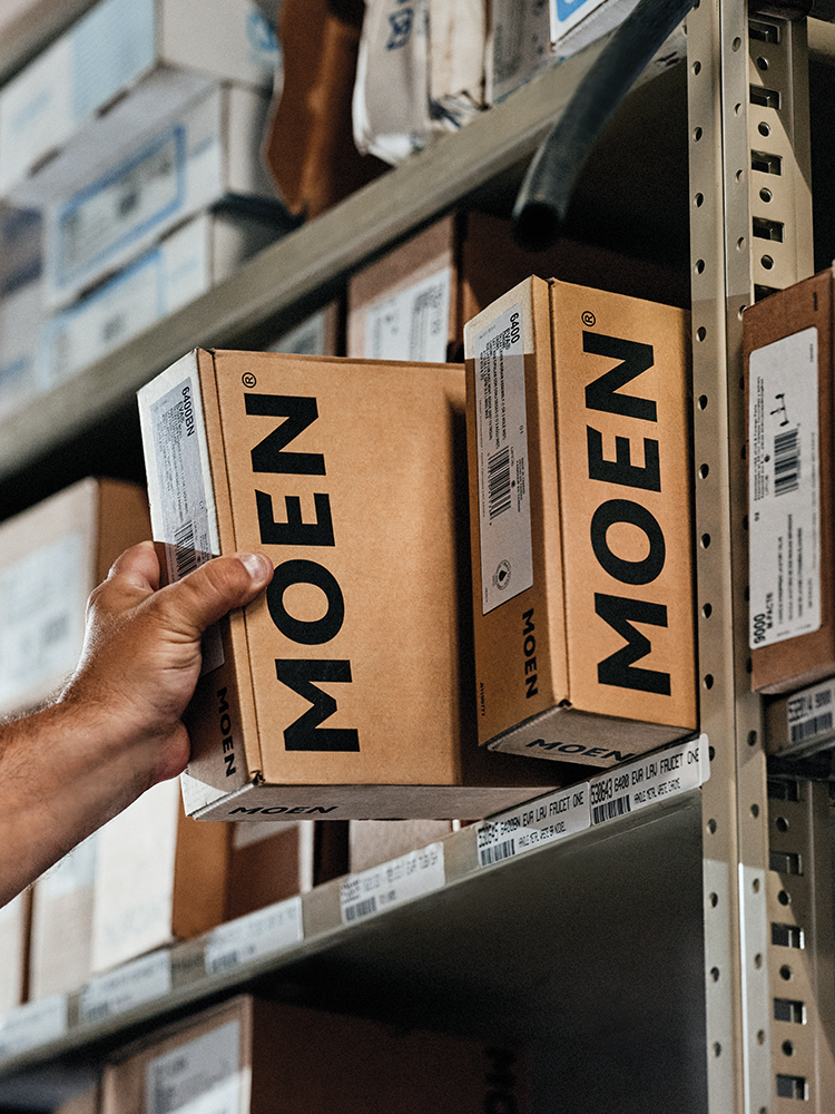 A hand holds a Moen product box next to another Moen box on a metal warehouse shelf, with other boxes visible in the background.