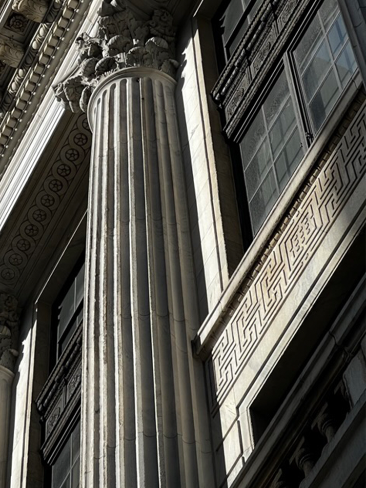 Close-up architectural detail of a large fluted stone column with ornate capitals and carved decorative molding beside dark window panes.