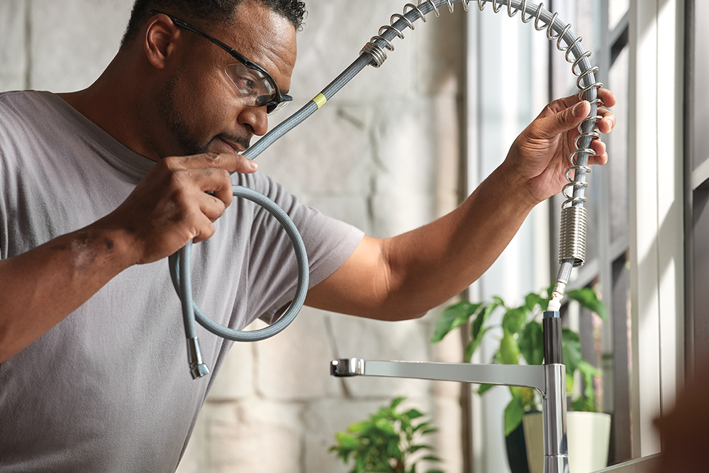 A man wearing glasses and a gray t-shirt is shown in a kitchen setting, demonstrating how to unscrew a flexible kitchen faucet hose.