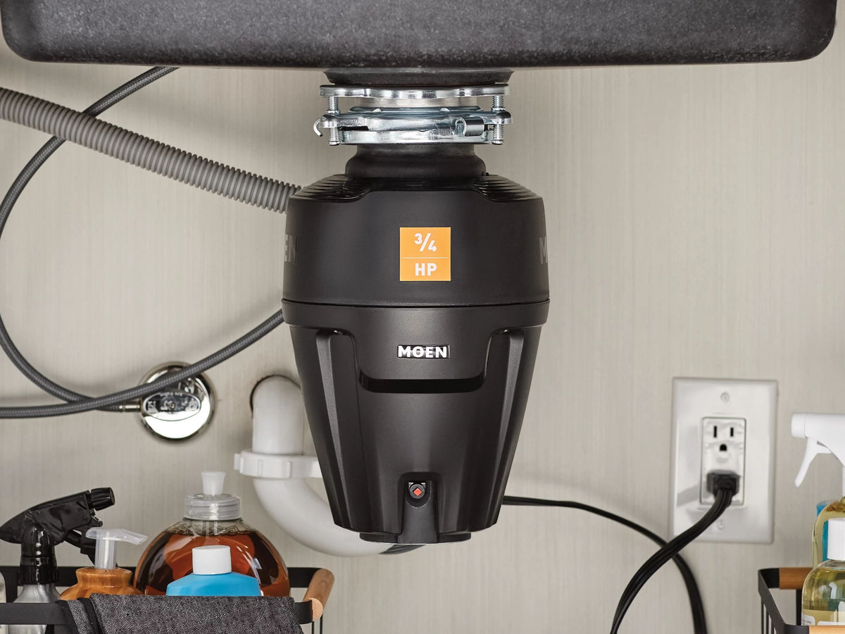 A black Moen garbage disposal under a sink, shown installed in a kitchen cabinet with visible plumbing, cords, and an outlet.