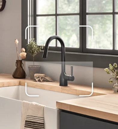 A matte black kitchen faucet installed on a light wood countertop in a farmhouse-style sink, shown in a real kitchen setting with a window, plants, and decorative vases.