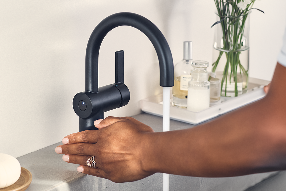 A matte black modern kitchen faucet with a curved gooseneck spout, shown with running water and a person's hand reaching under the spout in a bathroom or kitchen setting.