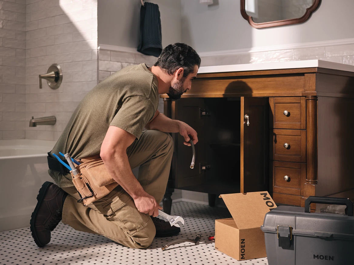 A male plumber or handyman kneels on a tiled bathroom floor next to a wooden vanity, working under a sink cabinet with tools in his waist belt and boxes branded MOEN on the floor.