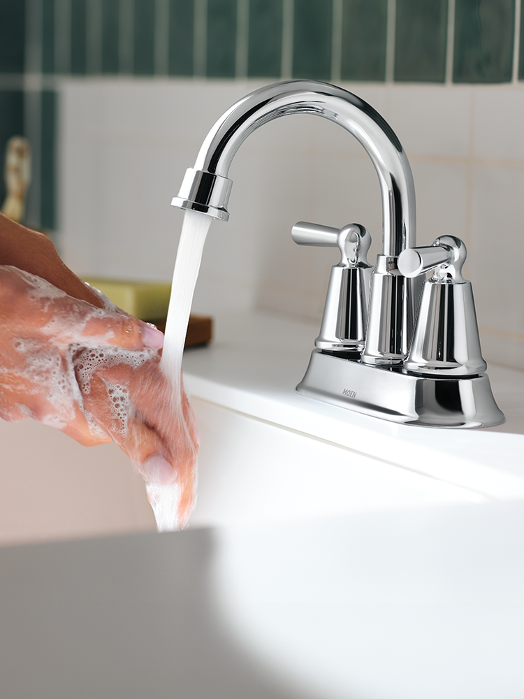 A polished chrome three-hole bathroom faucet with running water, shown installed in a white sink with a person's hand washing beneath it.