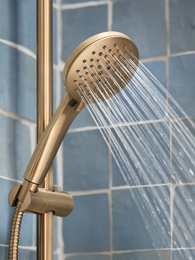A brushed gold shower head with water spraying from it, mounted on a matching gold shower rail against a blue tiled wall.