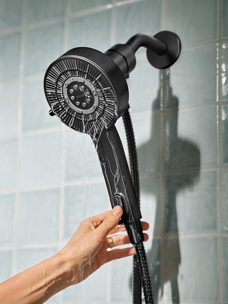 A matte black handheld shower head with a matching black hose, held by a hand against a light blue tiled wall with water running from the shower head.