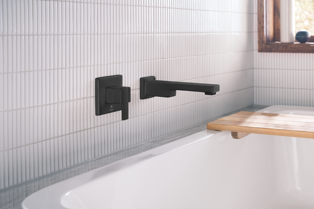 A matte black wall-mounted bathtub faucet and matching control handle installed on a white ribbed tile wall above a white bathtub, with a wooden bath mat visible in the corner.
