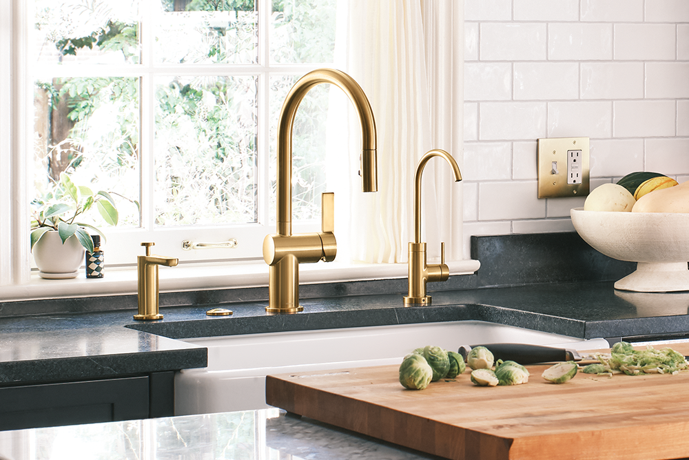 A modern kitchen scene featuring a brushed gold high-arc Cia faucet, a matching small side faucet, and a separate gold soap dispenser on a dark countertop. A white subway tile backsplash, a window with greenery outside, and a wooden cutting board with Brussels sprouts are also visible.