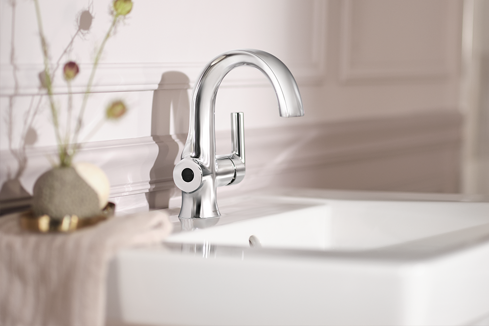 A polished chrome single-handle bathroom faucet mounted above a white sink, with a softly blurred beige wall and decorative dried stems in the background.