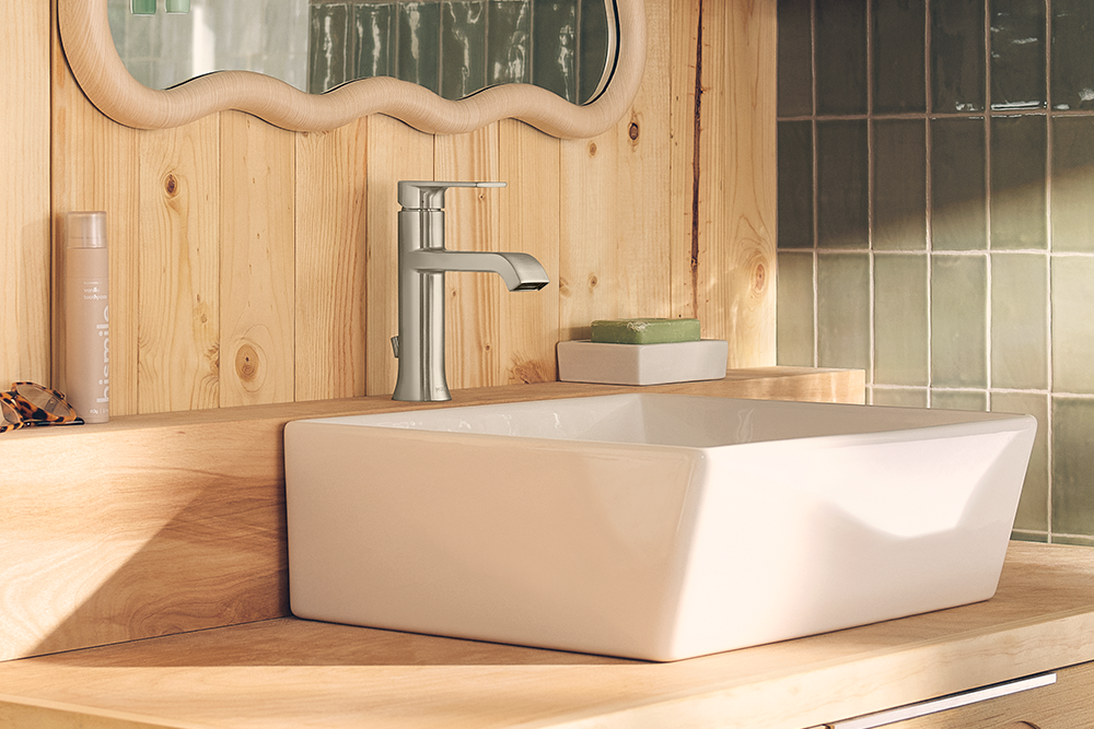 A modern white ceramic vessel sink with a matte finish, paired with a brushed silver faucet, set on a light wood vanity countertop in a bathroom with wood paneling and green-tiled walls.