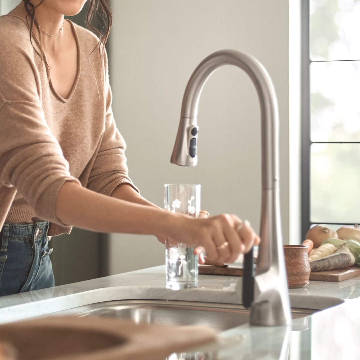 A woman in a beige sweater and blue jeans stands at a kitchen sink, filling a clear glass with water from a tall, curved matte silver faucet. A window and kitchen items are visible in the background.