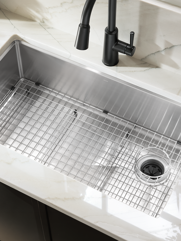 A stainless steel kitchen sink grid with a clear drain insert, installed in a white marble countertop with a black faucet.